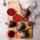 Four raw beets, including two halved pieces showing deep red interiors, arranged on a wooden cutting board with a striped cloth, a small jar of peppercorns, and a small bowl of red liquid. A round label reading 'PICKLING SPICE' is visible near the beets.