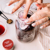 A close-up lifestyle shot of hands placing sliced red onions into a glass mason jar filled with dark redging liquid and ice, with a small bowl of red vinegar and a jar of Morton & Barrett Pickling Spice nearby on a light surface.