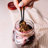 A close-up of a hand with light pink manicured nails adding a spoonful of whole spices and peppercorns into a clear glass mason jar filled with dark red wine, onions, and garlic. A striped kitchen towel and a red lid are softly blurred in the background.