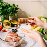 A person's hands pour a spice blend from a clear glass shaker bottle into a metal measuring spoon over raw chicken pieces in a glass bowl, set against a kitchen backdrop with fresh vegetables.