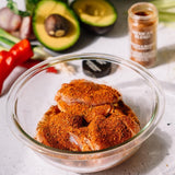 Seasoned chicken pieces in a clear glass bowl, with a jar of Mexican Blend seasoning and fresh ingredients like avocado, red pepper, and garlic in the background.