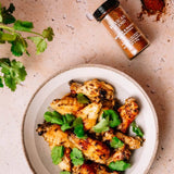 Grilled chicken wings garnished with fresh cilantro in a ceramic bowl, with a bottle of Mexican Blend seasoning and cilantro sprigs on a textured beige surface.