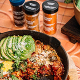 A cast iron skillet filled with cooked food topped with sliced avocado, cilantro, and cheese, with three spice jars in the background on a terracotta surface.