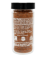 A clear plastic spice jar filled with brown seasoning blend, topped with a black lid, shown against a solid black background. The back label is visible, showing the brand Asa & Bassett Spices and product details.