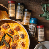 A styled flat-lay of Morton & Bailey restaurant spices on a rustic wooden table, including Garlic Powder, Italian Seasoning, Black Pepper, Onion Powder, and Morton & Bailey Anchovy, arranged around a pan of baked meatballs and cheese.