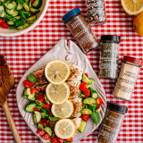 A styled flat-lay of a grilled chicken salad topped with lemon slices, cucumber, and tomato, surrounded by several spice jars on a red-and-white checkered tablecloth.