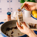 A person holds a glass jar of Italian Seasoning in San Francisco over a blue pan, with a wooden spoon lifting seasoning from the jar. Another seasoning bottle and lemons are visible in a kitchen setting.