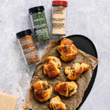 Four spice and seasoning bottles arranged on a light stone surface beside a black baking tray holding several golden baked rolls on parchment paper. Visible labels include Parsley, Garlic Powder, Italian Seasoning, and Morton & Bassett Italian Seasoning.