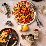 Overhead flat-lay of a seafood pasta dish with shrimp, cherry tomatoes, and lemon slices on a plate, surrounded by seasoning bottles, forks, and a glass on a light stone surface.