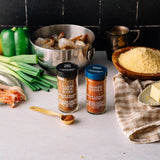 Two spice jars from The Spice Store displayed on a light countertop with cooking ingredients and a pot of shrimp around them. The jars are labeled 'Cajun Spice Blend' with a black lid and 'Smoked Paprika' with a blue lid.