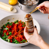 A person holds a small spice jar labeled 'Cajun Spice Blend' over a pan of chopped vegetables, with a butter dish and pot blurred in the background.