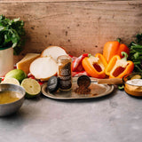 A jar of Calm Spice Blend seasoning sits on a silver tray surrounded by fresh ingredients including bell peppers, onions, limes, and herbs on a rustic wooden surface.