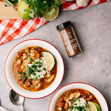Two bowls of chicken and bean chili topped with sour cream, chopped green onions, and lime wedges, arranged on a gray surface with a bottle of Chili Spice Blend, a red-and-white check cloth, lime slices, garlic, and a block of cheese.