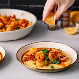A plated shrimp and grits dish on a light gray surface, with a hand squeezing lemon juice over the food. A large bowl of shrimp is blurred in the background, along with bottles and additional lemon wedges.