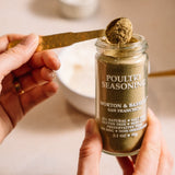 A person holds a glass jar of Morton & Bassett San Francisco Poultry Seasoning while another hand uses a gold spoon to scoop some of the seasoning from the jar.