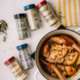 A styled flat-lay of five spice jars arranged around a white enameled cast-iron pot filled with cooked chicken and herbs on a light surface. The jars include Thyme, Chicken Seasoning, Ground Ginger, Garlic Powder, and Ground Pepper.