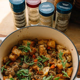 A close-up of a prepared stuffing or bread mixture in a cream-colored Dutch oven, garnished with fresh herbs, with several spice jars standing behind it on a wooden surface.