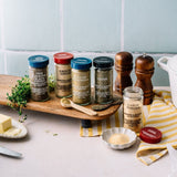Four glass spice jars with blue, red, and dark lids are arranged on a wooden board with peppercorns, a small dish of garlic powder, herbs, and a striped cloth in a kitchen setting.
