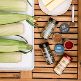 A styled flat-lay of seasoning bottles and jars alongside raw ingredients on a wooden slatted board and white platter. Visible items include several spice bottles labeled Garlic Powder, Barbecue Rub, Herb Rub, and Salted Paprika, along with ears of corn, butter, and a measuring spoon.
