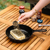 A hand sprinkles seasoning from a metal measuring spoon onto a rectangular bar of butter resting in a black cast-iron skillet on a wooden slatted table. Several spice jars are visible in the background.