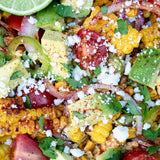 Close-up of a colorful Mexican corn salad with grilled corn kernels, diced avocado, red tomato, onion, cilantro, and crumbled white cheese.