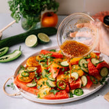 A hand holds a small glass bowl and pours a reddish-brown dressing over a colorful watermelon salad on a white oval platter. Slices of watermelon, cucumber, lime, jalapeño, and mixed tomatoes are visible, with additional cucumbers, limes, herbs, and an orange in the background on a light countertop.
