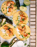 Four shrimp tacos arranged on a light blue tray with limes, grilled peppers, cilantro, and a small bowl of white sauce. The photo is taken from above on a wooden slatted surface.