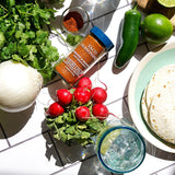 A styled flat-lay of taco seasoning in a clear spice jar with a blue lid, surrounded by fresh ingredients including cilantro, radishes, jalapeño, lime, onion, tortillas, and a glass of iced water on a white tiled surface.