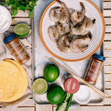 Overhead flat-lay of shrimp on a white plate with taco seasoning bottles, limes, tortillas, chopsticks, and a small bowl on a rustic wooden surface.