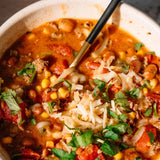 A close-up shot of a hearty vegetable chili soup in a speckled cream-colored bowl, topped with shredded cheese and fresh cilantro, with a black-handled spoon resting inside.