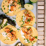 Four shrimp and corn tacos arranged on a metal tray with lime wedges, sliced zucchini, and chopped cilantro. The tray is photographed from above on a light wood slatted surface.