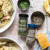 Two bottles of Morton & Bassett Italian Parsley and Coarse Ground Black Pepper seasoning arranged on a rustic white wooden table with food props including risotto, parsley, lemon, and a grating board.