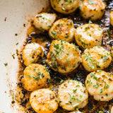 Pan-seared scallops topped with chopped fresh parsley and herbs, cooking in a pan with visible oil and browned edges.