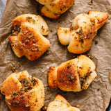 Several golden-brown baked rolls or bread twists topped with herbs and seasoning, arranged on crumpled brown parchment paper.