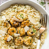 A close-up overhead shot of a white bowl filled with creamy risotto and scallops topped with herbs and shaved cheese, with a gold-toned fork on the right side.