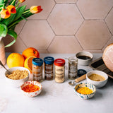 A styled kitchen scene showing several spice jars and bowls of ground spices, with whole and sliced oranges, tulips in a vase, and a tiled wall in the background.