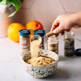 A hand with white nail polish pours a scoop of ground ginger powder from a metal measuring cup into a speckled white bowl. In the background, three spice jars (Ground Ginger, Garlic Powder, and Black Pepper) and oranges sit on a light marble countertop.