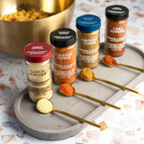 Four jars of Morton & Bastey seasoning spices arranged on a gray tray with gold spoons holding the powder, plus a gold bowl in the background on a light stone surface.