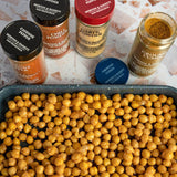 Spices including cayenne pepper, chili powder, garlic powder, and ground cumin above a tray of chickpeas.