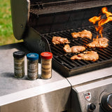 Three spice seasoning bottles labeled Rosemary, Fine Ground Pepper, and Garlic Powder sit on the side of a stainless steel grill next to chicken cooking over an open flame.