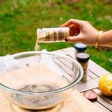A hand pours garlic powder from a clear plastic shaker bottle into a large glass bowl on a wooden table, with a green grassy background.