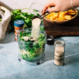 A hand pours onion powder from a metal scoop into a food processor containing chopped green vegetables. A glass jar of onion powder sits on the counter next to the processor, with a bottle of garlic powder and a bowl of tortilla chips in the background.