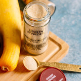 A glass jar of Morton & Bassett onion powder sits on a wooden tray with a gold measuring spoon, a red-lidded jar, and a yellow squash on a blue textured background.