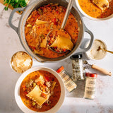 Overhead flat-lay of a hearty pasta soup dish with a large pot and two bowls of orange-red broth, pasta, and ground meat, surrounded by spice jars and small bowls on a light stone surface.
