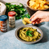 A bowl of green guacamole dip with a cilantro garnish, set on a blue-gray surface. A hand holds a tortilla chip dipped in guacamole.