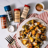 A styled flat-lay of roasted Brussels sprouts on a white plate with shaved cheese, surrounded by four seasoning bottles labeled Smoked Paprika, Black Pepper, Garlic Powder, and Onion Powder, along with a honey dipper and a fork on a white marble surface.