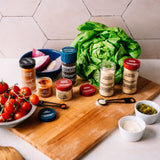 A styled flat-lay of gourmet seasoning blends and fresh ingredients on a wooden cutting board, featuring bottles and jars of spices alongside tomatoes, onion, and a head of lettuce.