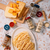 An overhead flat-lay of pasta seasoning spices arranged around a plate of spaghetti and cheese on a white marble surface. Visible spice jars include garlic powder, onion powder, dried garlic, dried onion, and red pepper flakes, with cheese wedges and cubed mozzarella on a wooden board.