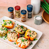 Six jars of Gourmet Seasonings are arranged on a pink countertop behind a white platter of stuffed bell peppers topped with melted cheese and chopped green onions.