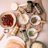 An overhead flat-lay of Italian seasoning blends and spices arranged on a striped brown and beige cloth with bowls of ingredients, including sun-dried tomatoes, garlic powder, dried herbs, parsley, and shredded cheese, on a light beige surface.